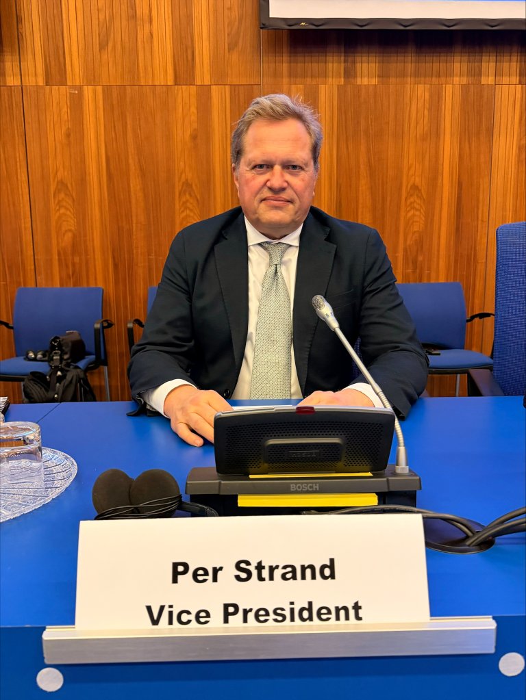 Mann sitter ved et konferansebord med mikrofon; foran ham står en navneplate med teksten «Per Strand, Vice President», i et formelt møterom med blå stoler og trepanelte vegger.