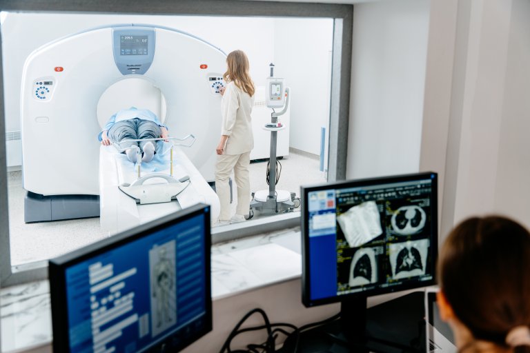En pasient ligger i en CT-maskin mens en operatør styrer maskinen. Skjermer med CT-bilder i forgrunnen.