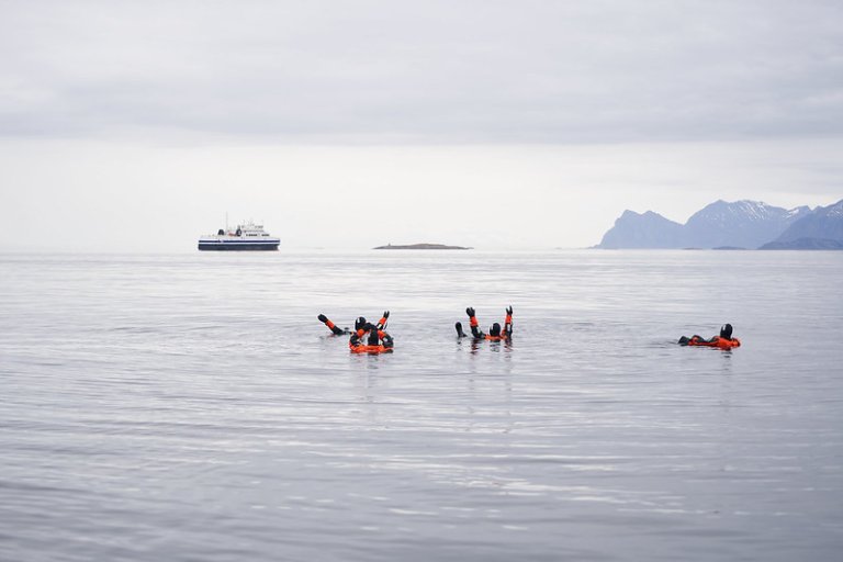 Et skip og fire personer med overlevelsesdrakt i vannet.