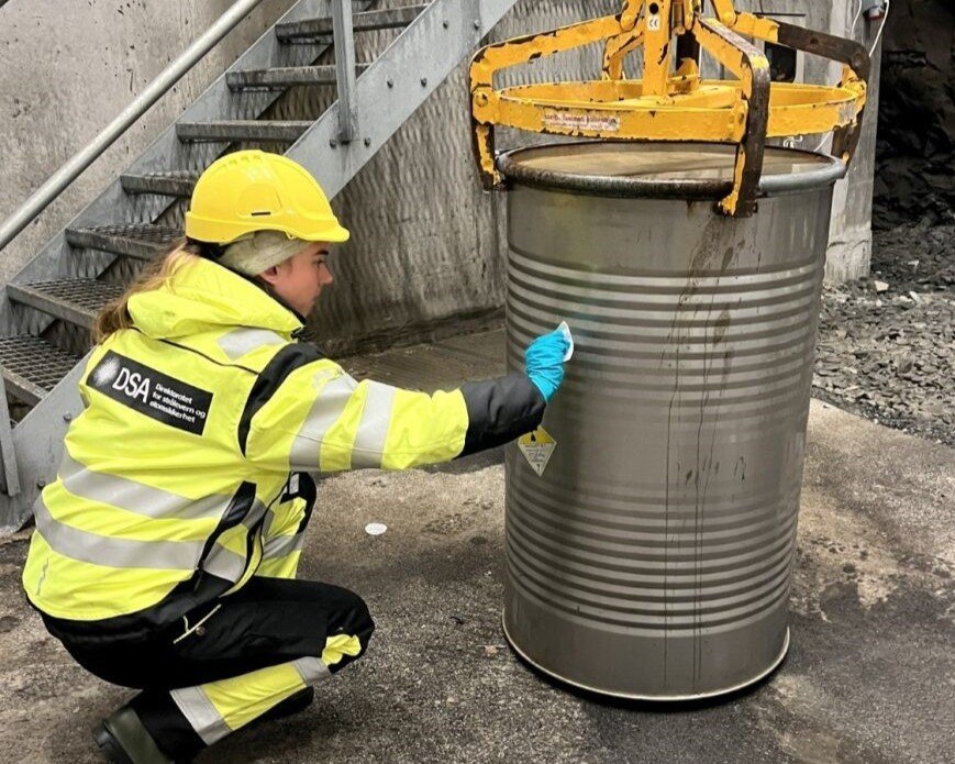 Ung kvinne i gul vernejakke og hjelm stryker et papir mot en metalltønne merket med symbol for radioaktivt innhold.