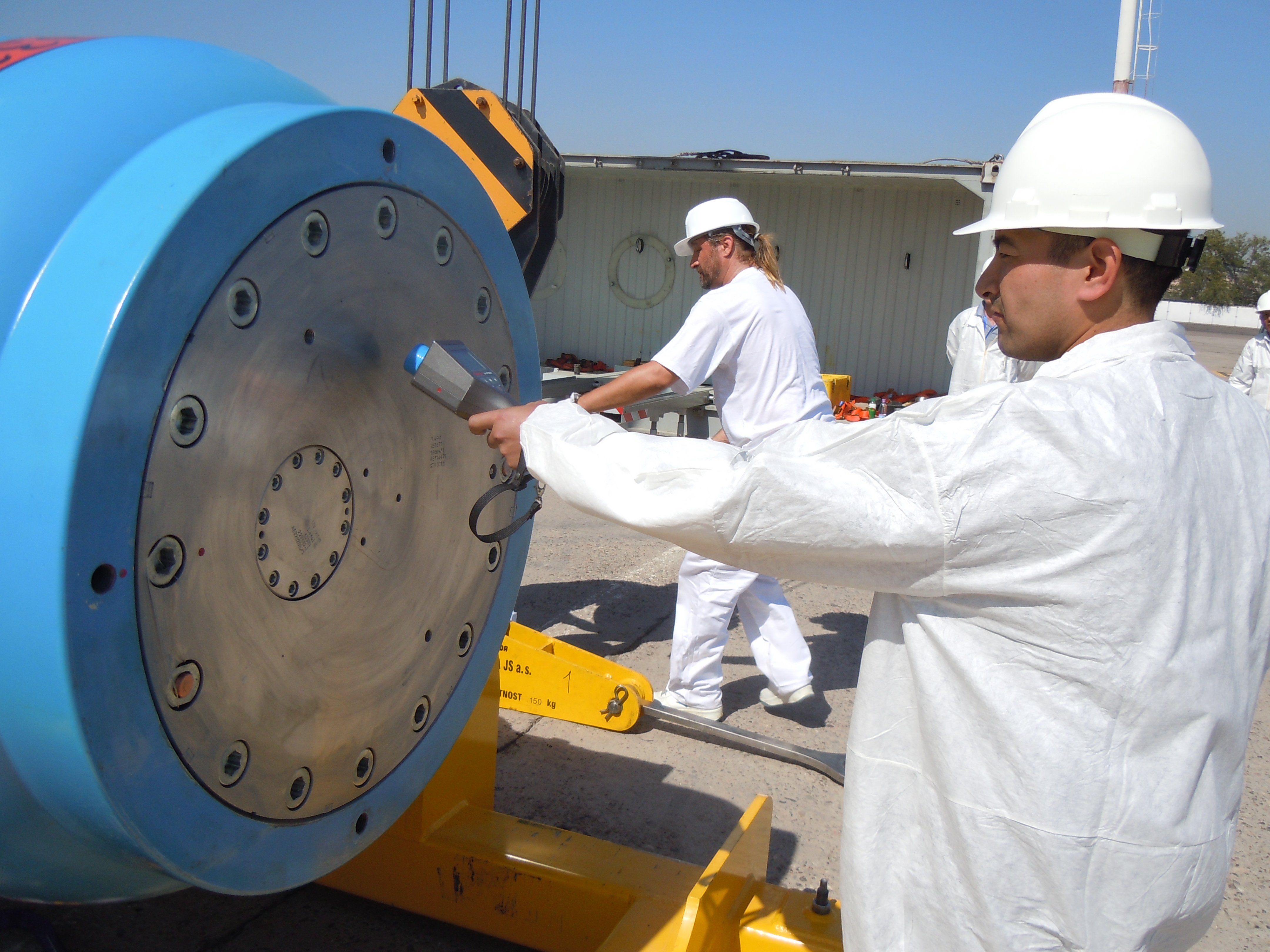 Mann i kvit frakk og hjelm held eit måleinstrument mot ein stor gjenstand i metall.
